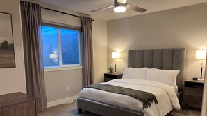 A bedroom with a bed, two bedside tables with lamps, a ceiling fan, a window with curtains, and a rug. - Cozy Cascade Nook - 2 bedroom basement apartment (OREM)