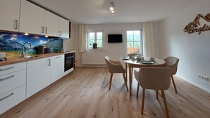 A modern kitchen with white cabinets, a wooden countertop, and a painting of a mountain landscape. There is a dining area with a table and chairs, a window with curtains, and a flat-screen TV mounted on the wall. - Ferienwohnung 4 mit Balkon (Berchtesgaden)