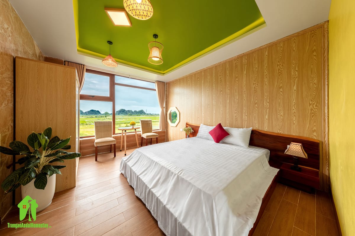 Slaapkamer met een groot bed, een houten hoofdbord, een nachtkastje, een kamerplant, een raam met uitzicht op de omgeving en een plafond met groene en witte panelen.