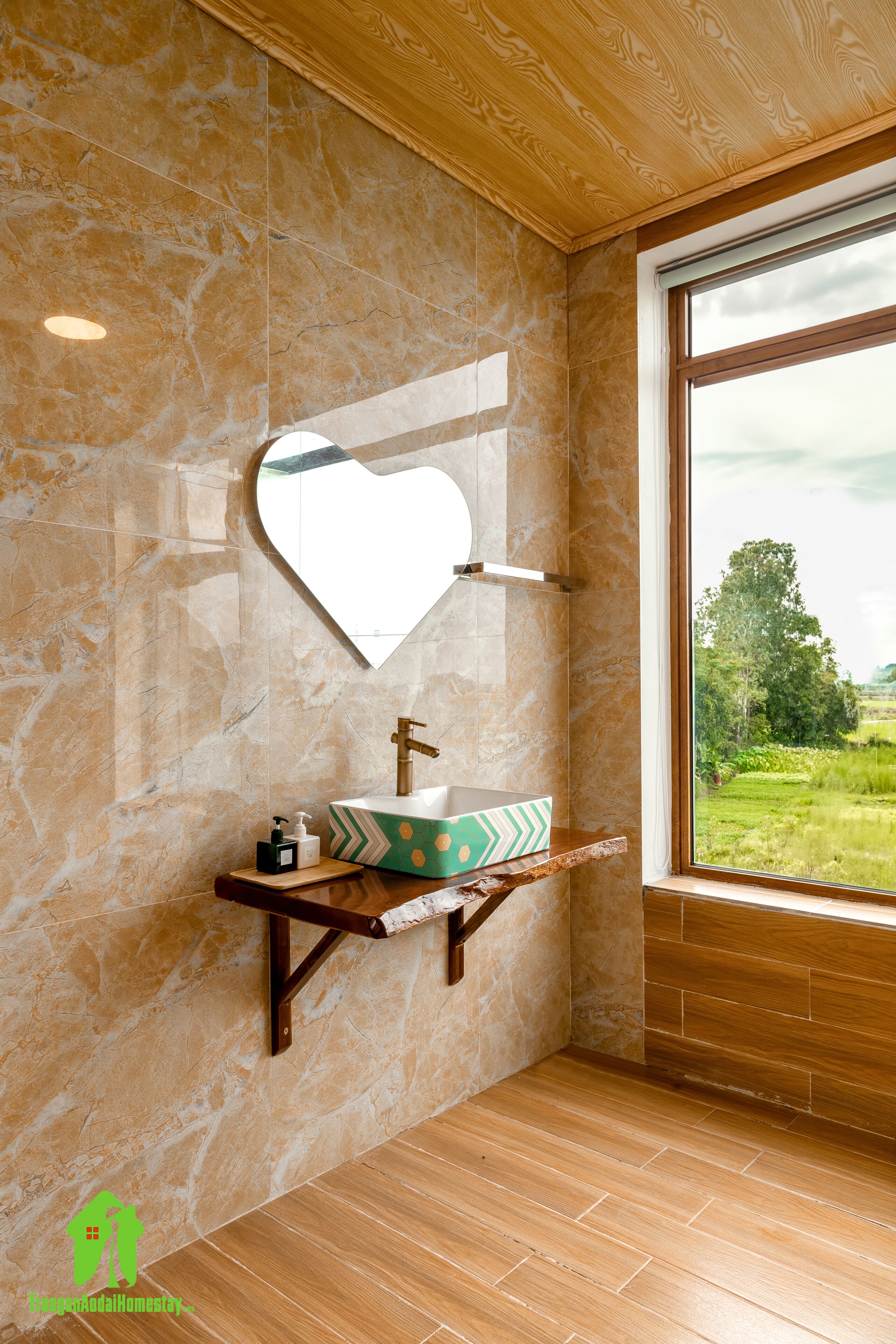Un baño con pared de mármol, tocador de madera, espejo con forma de corazón y ventana con vistas a zonas verdes.