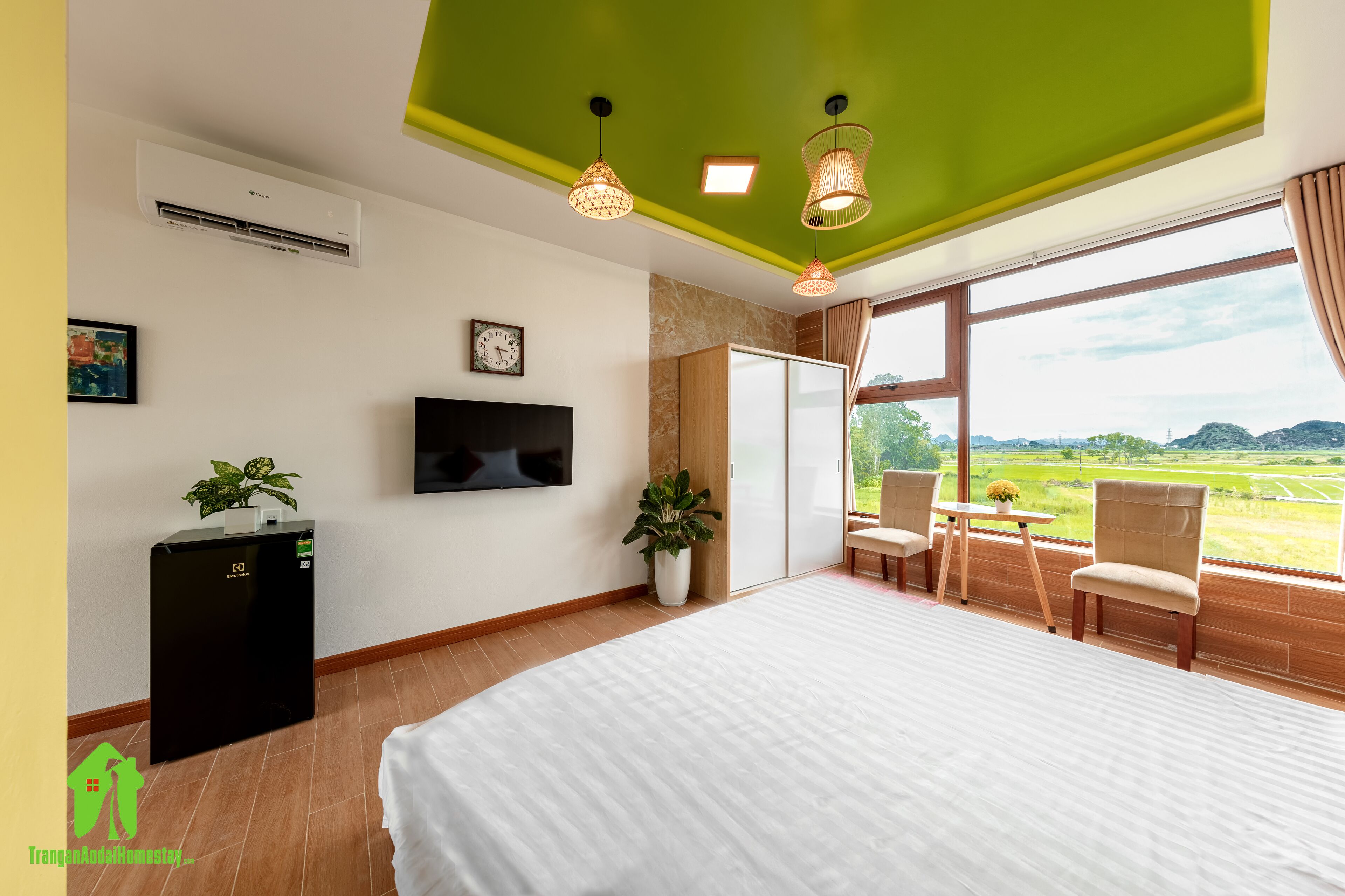 Habitación de hotel con una cama grande, un televisor de pantalla plana, un refrigerador pequeño, una planta en maceta, una mesa de comedor con sillas y vistas a un paisaje verde a través de amplias ventanas.