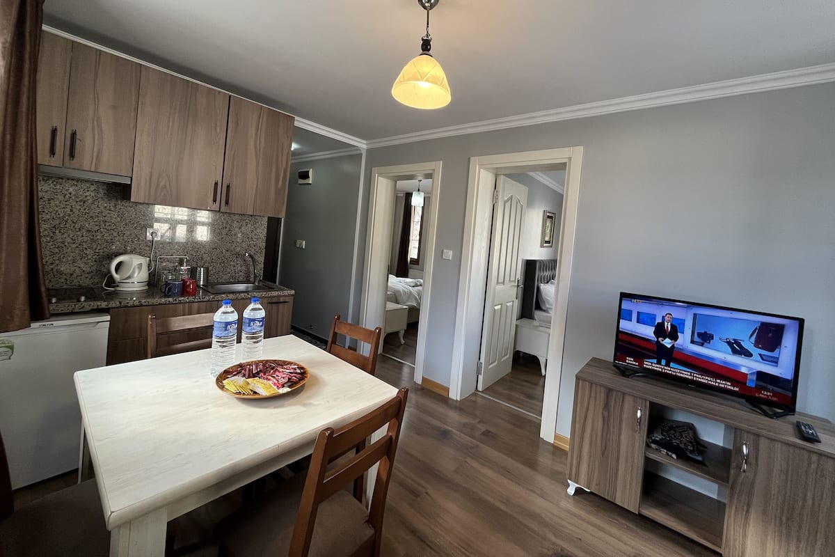 A modern kitchen with wooden cabinets, a dining table with chairs, a television, and a small cabinet with a remote control.