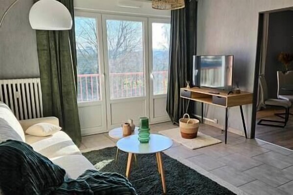 Una sala de estar moderna con un sofá, una mesa de centro, un televisor sobre un mueble bajo y vistas al exterior a través de amplios ventanales.