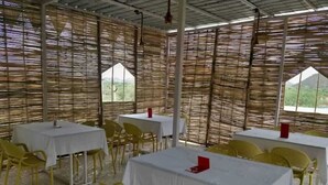 An outdoor dining area with tables and chairs, covered by a bamboo roof, and large windows offering a view of the landscape.