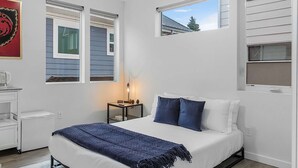A bedroom with a bed, a nightstand, a lamp, and a window with a view of a house. - Balerion's Shadowed Suite (Seattle)
