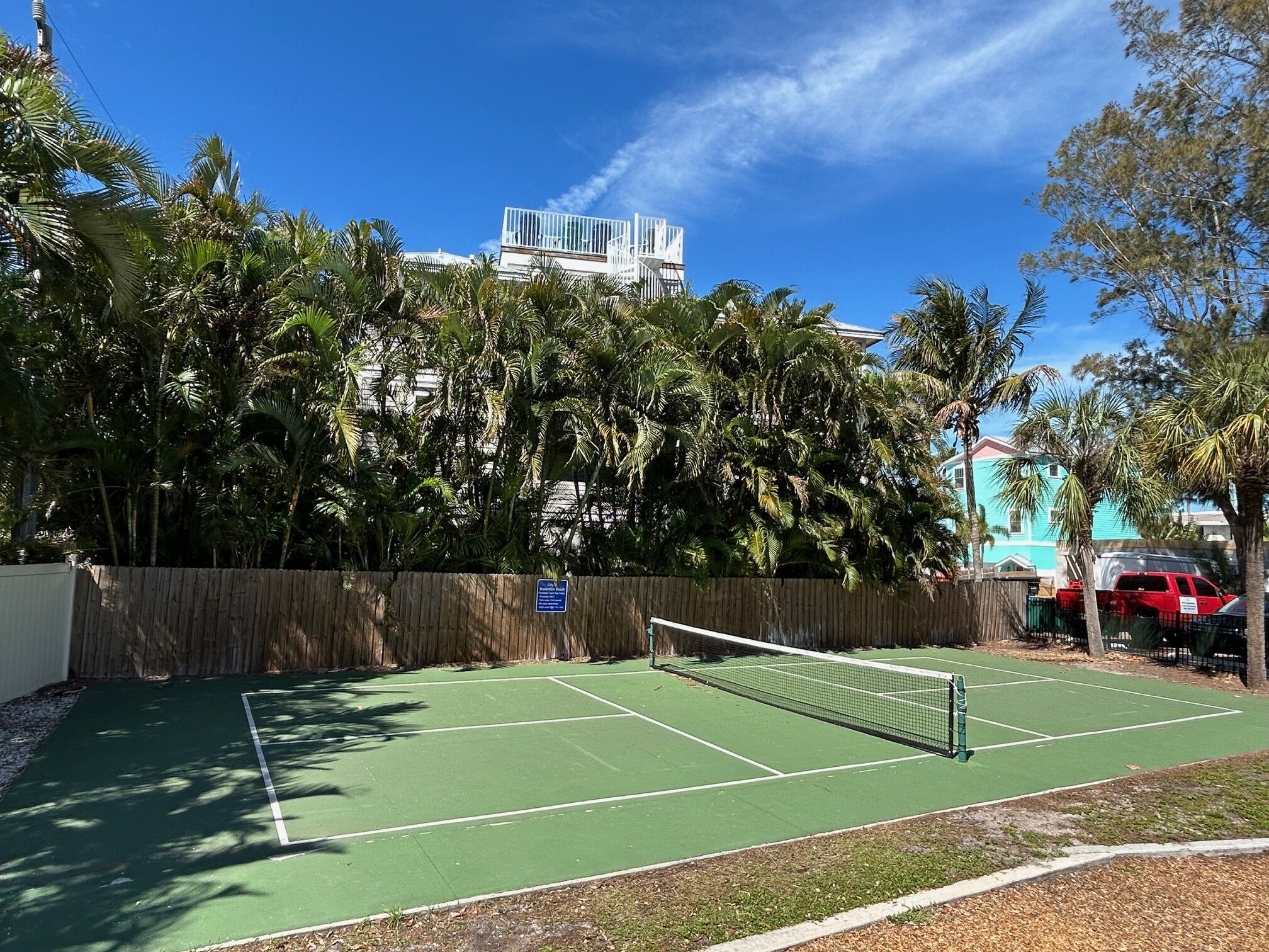 Un court de tennis entouré d’une clôture en bois, avec des palmiers en arrière-plan.