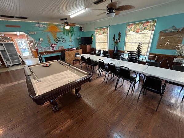 A pool table in a room with a long table and chairs, a ceiling fan, and a wall with a tropical mural. - Bridge Street Pier Beach House + Dog-friendly (Bradenton Beach)