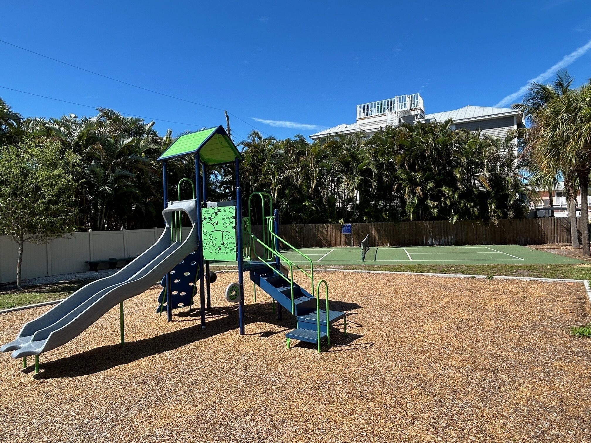 A playground with a slide and climbing structures, surrounded by trees and a tennis court in the background.