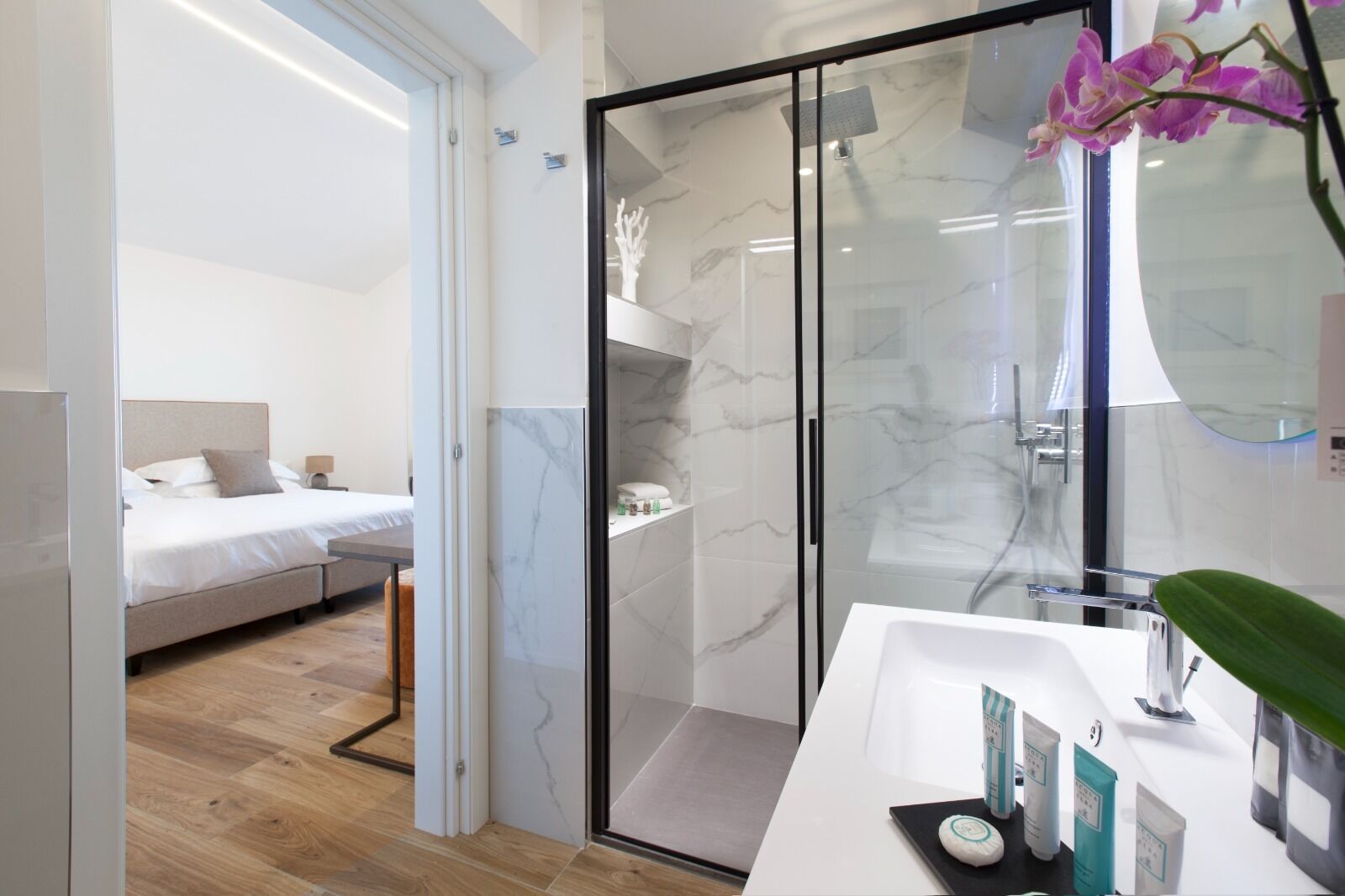 A modern bathroom with a glass shower enclosure, a white sink, and a bed visible through an open door.