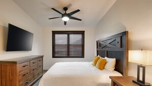 A bedroom with a bed, a wooden dresser, a ceiling fan, a TV mounted on the wall, and a window with blinds.