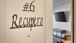 A room with a #6 Recupero sign, a flat-screen TV mounted on the wall, a wooden dining table, and a grey cabinet.