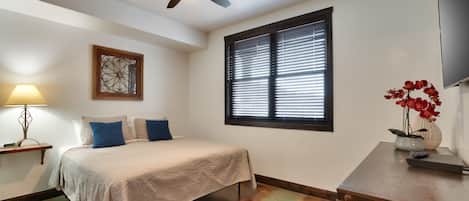 A bedroom with a bed, a ceiling fan, a window with blinds, a wall-mounted TV, and a bedside table with a lamp and a vase of flowers.