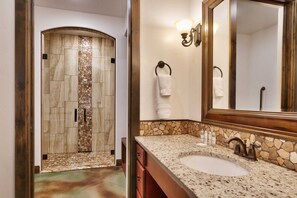 Une salle de bain avec une douche carrelée de mosaïques en pierre, un lavabo avec un plan de travail en granit et un grand miroir.