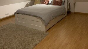 Une chambre à coucher avec un lit, un ventilateur de plafond, un luminaire, une petite fenêtre et un plancher en bois.