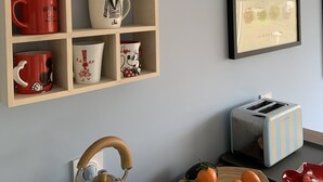 Une cuisine avec une étagère en bois où sont posées différentes tasses, une bouilloire, un grille-pain et une assiette remplie d’oranges.