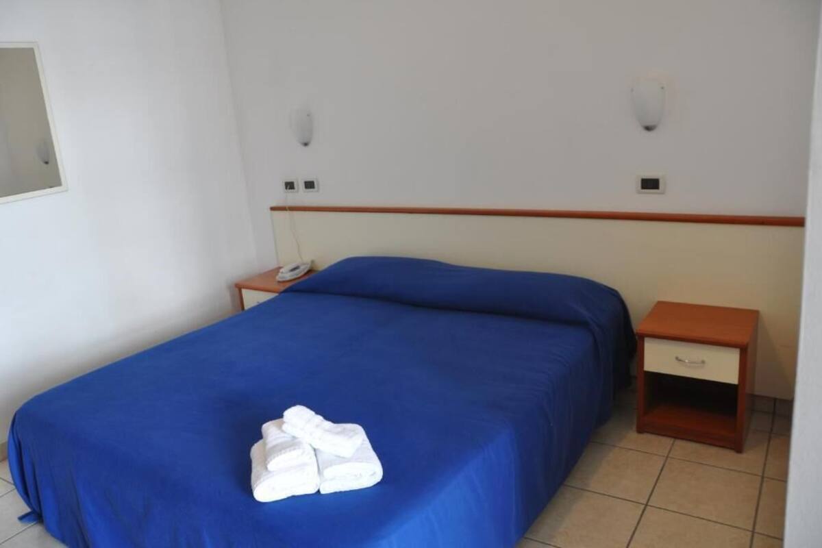 Una cama individual con colcha azul y toallas blancas, una mesita de noche de madera con cajón, y un teléfono instalado en la pared.