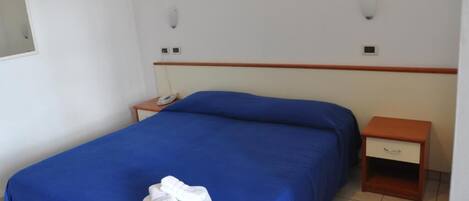 Una cama individual con colcha azul y toallas blancas, una mesita de noche de madera con cajón, y un teléfono instalado en la pared.
