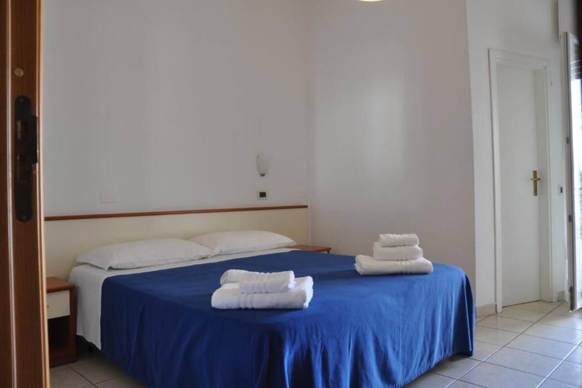 Una habitación de hotel con una cama, dos toallas blancas, una colcha azul, un cabecero de madera, una pared blanca y una puerta.