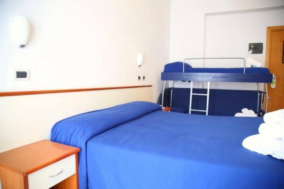 Una cama litera con colchón azul, una mesita de noche de madera con cajón, y una pared blanca con interruptor de luz y pequeño enchufe.