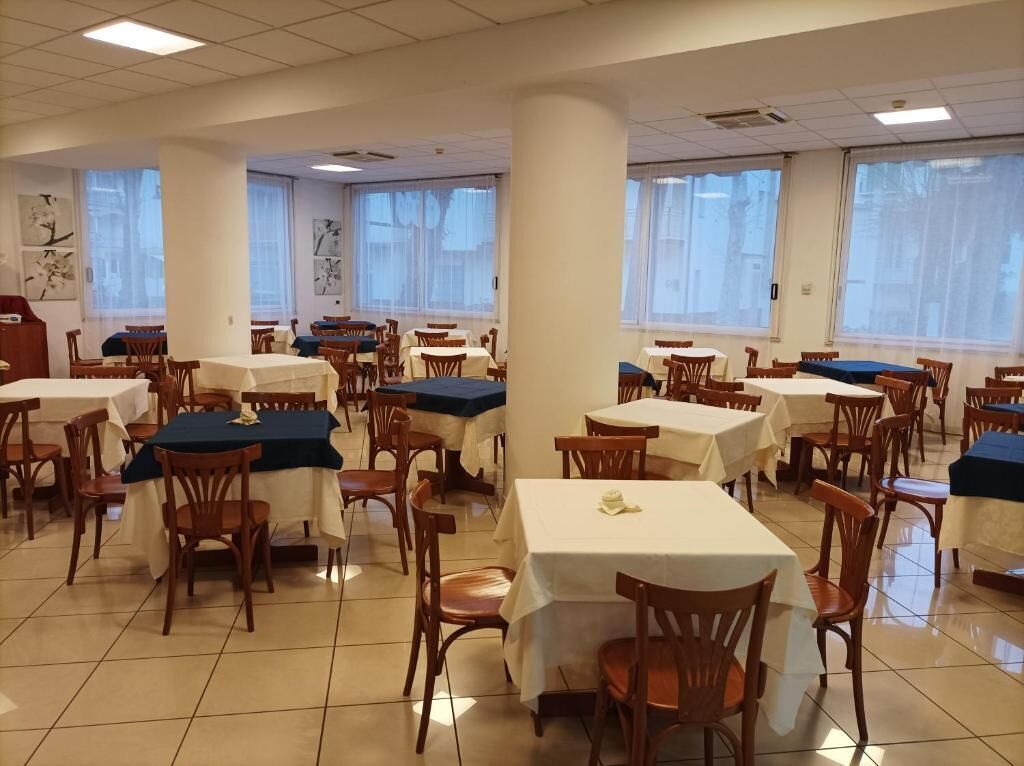 Spiseområde med flere borde og stole, store vinduer med lette gardiner, og et flisegulv.