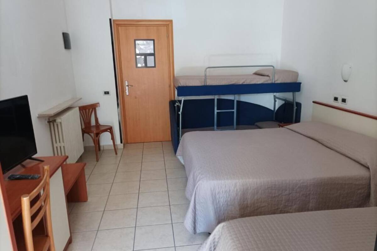 Habitación de hotel con literas, un escritorio con silla, televisor y una puerta con ventana pequeña.
