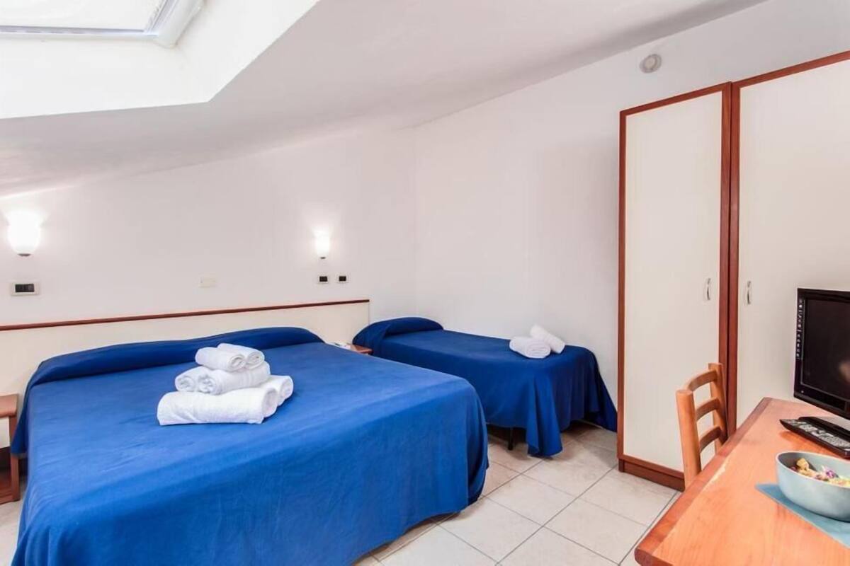 Habitación de hotel con dos camas, cada una con cubrecama azul y toallas blancas. Hay una mesita de madera con un bol y un mando a distancia.