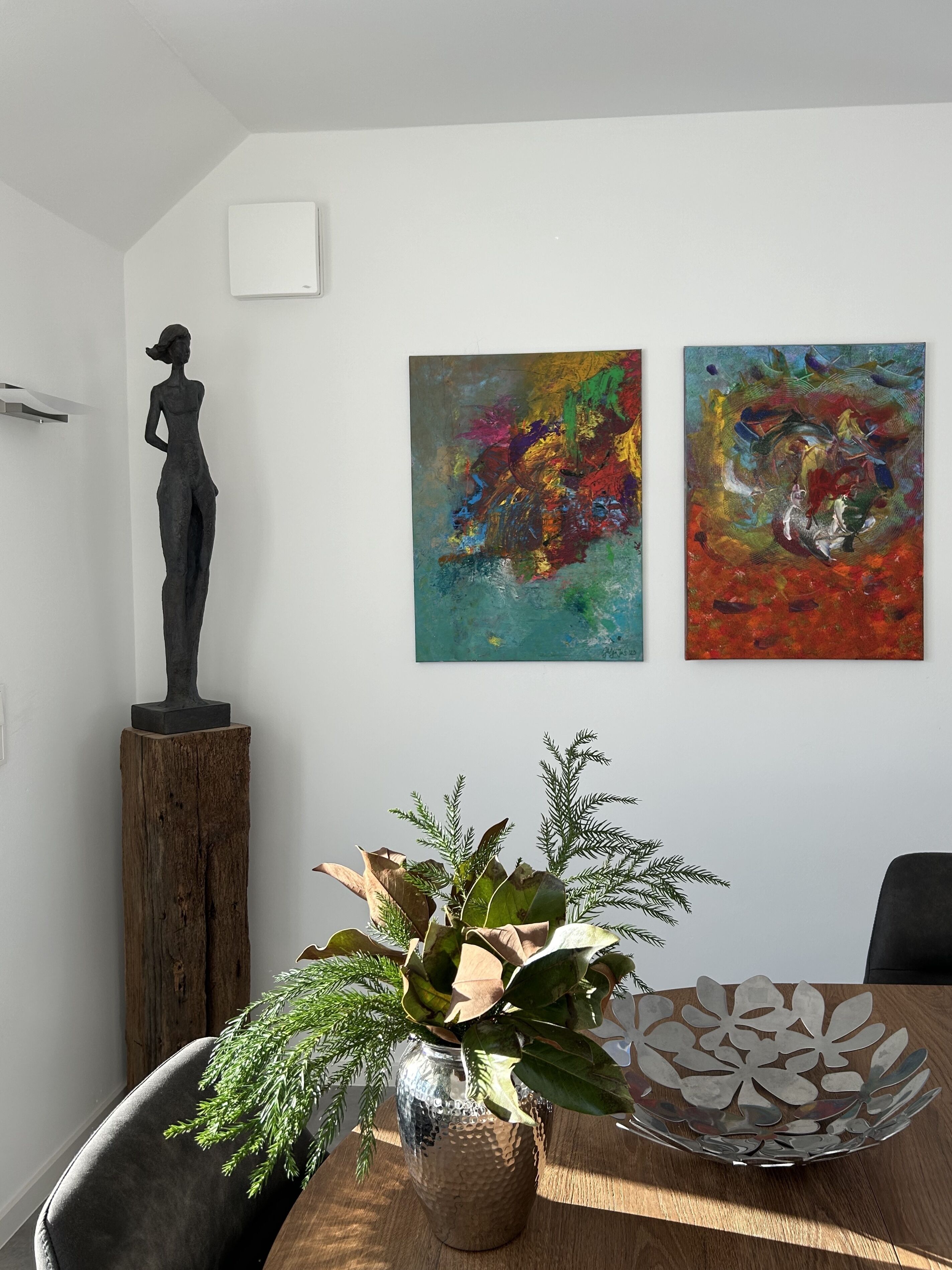 Ein Essbereich mit einem Holztisch, einer Metallschüssel, einer Topfpflanze und einer Statue auf einem Holzfuß. An der Wand befinden sich zwei abstrakte Gemälde.