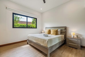 A bedroom with a large window, a bed with pillows, a bedside table with a lamp, and a nightstand.