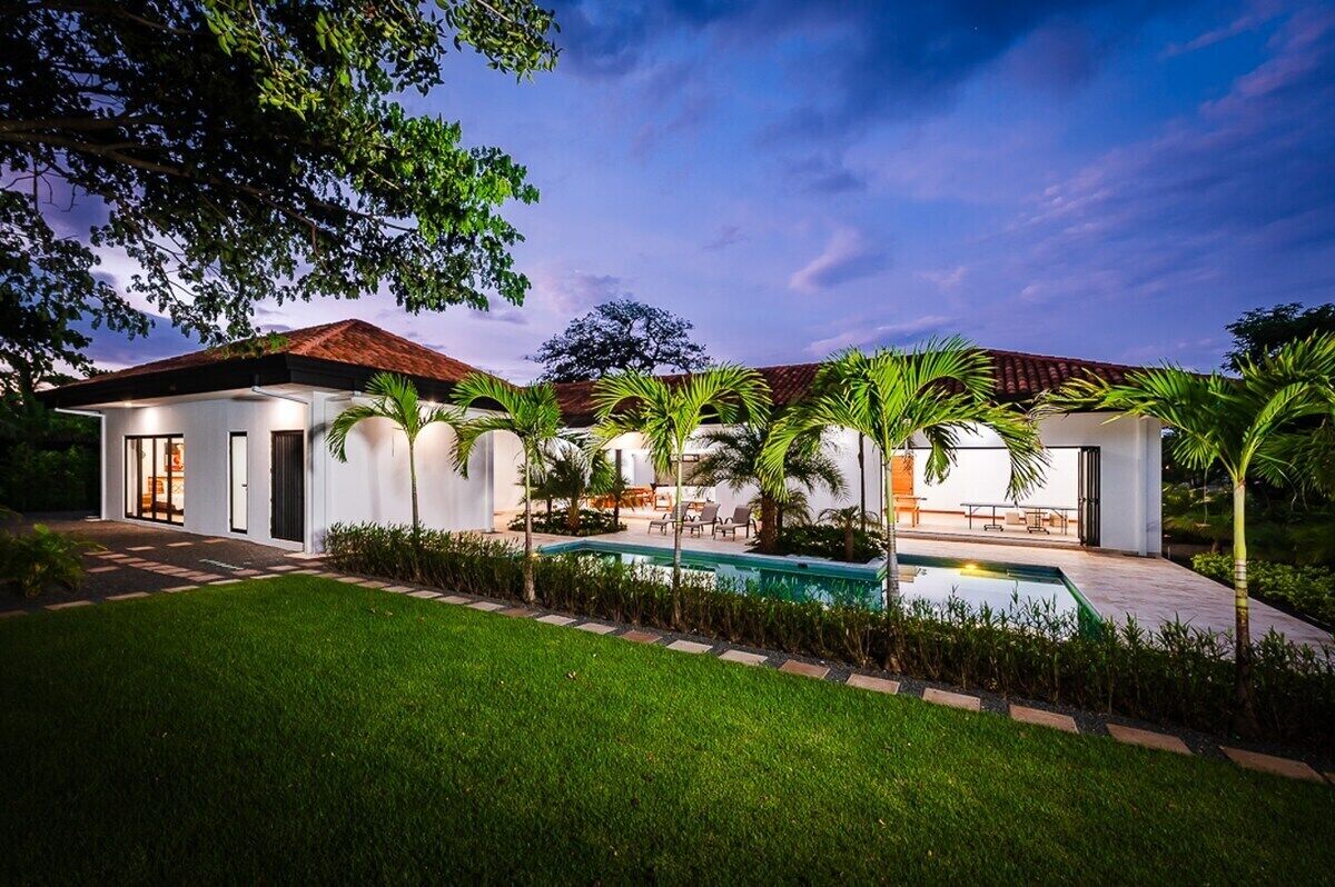 Uma casa moderna com piscina, cercada por gramado bem cuidado e palmeiras.