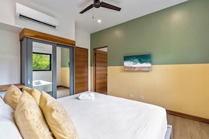 A bedroom with a bed, a ceiling fan, an air conditioning unit, a painting, and a window with a view of greenery.