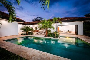 A swimming pool with a patio area, palm trees, and a modern building with a tiled roof. - Villa OSU (Reserva de Golf, Hacienda Pinilla)