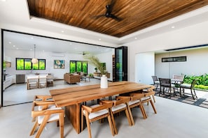 A modern kitchen with a wooden ceiling, a dining area with a large wooden table and chairs, and a view of greenery outside.