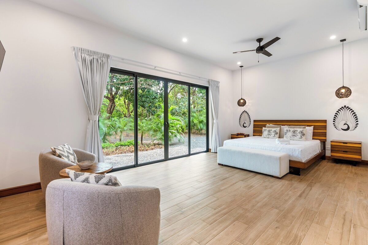 Un dormitorio moderno con una cama grande, un sofá, un ventilador de techo y vistas a la vegetación a través de las ventanas.