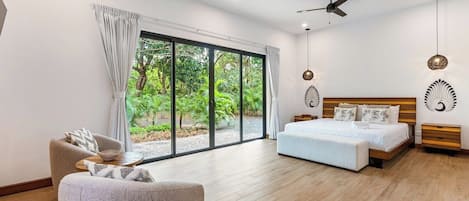 Une chambre moderne avec un grand lit, un canapé, un ventilateur de plafond et une vue sur la verdure par les fenêtres.