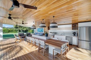 Area cucina esterna con terrazza in legno, tavolo da pranzo, sedie, ventilatori a soffitto e vista sul verde.