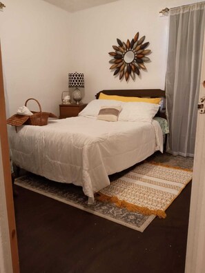 A neatly made bed with a white comforter and pillows, a decorative mirror on the wall, a small bedside table with a lamp, and a wicker basket. - Super Comfy Room at a Great Location  (Detroit)