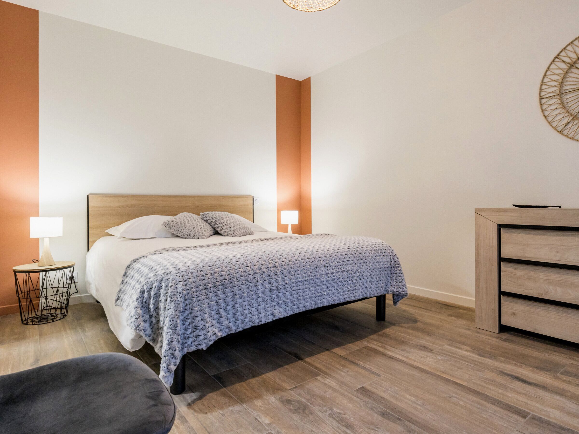 Un dormitorio moderno con cabecera de madera, colcha estampada de color gris, dos lámparas de noche, cómoda de madera y un sillón gris.