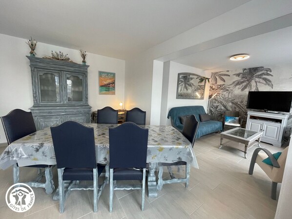 A dining area with a table and chairs, a cabinet, a sofa, a TV, and a wall with a tropical mural.