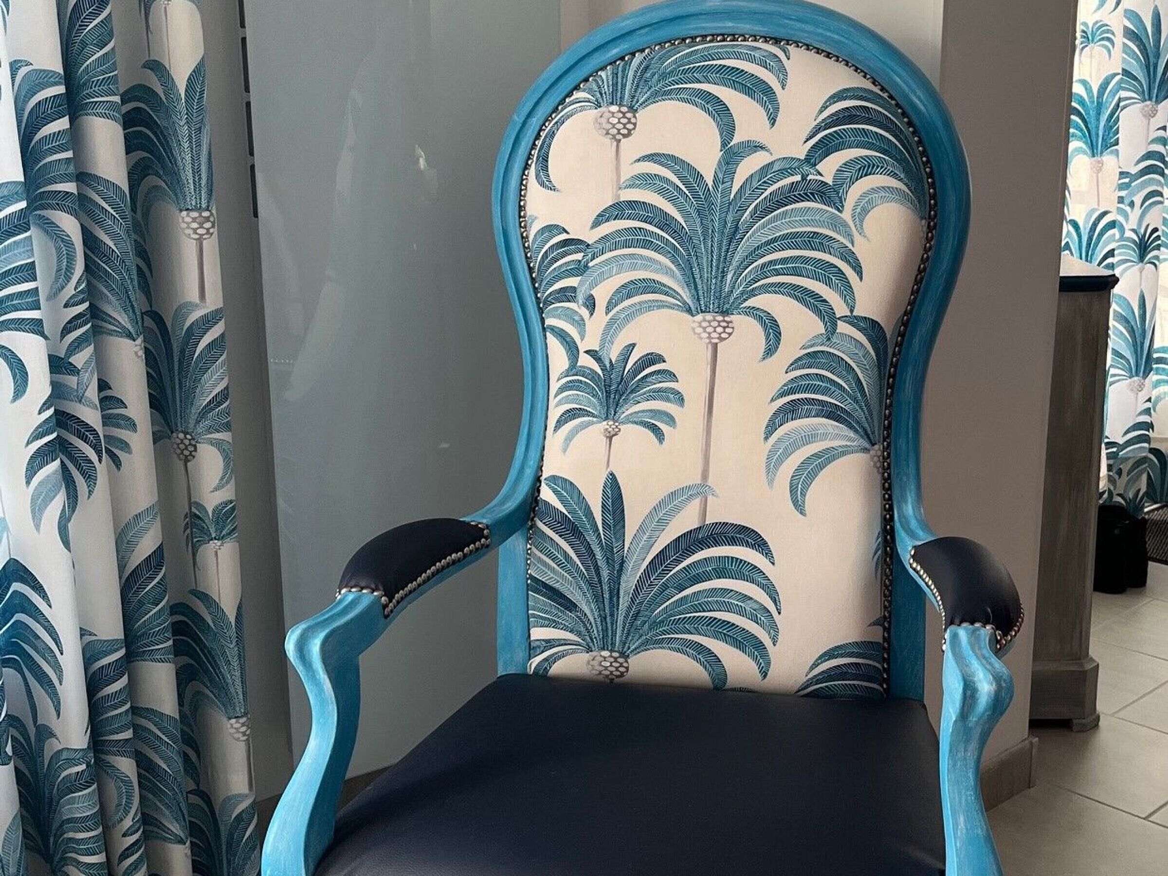 Une chaise dont l’habillage est orné d’un motif floral et dont l’armature est bleue, placée dans une pièce où les rideaux présentent un motif similaire.