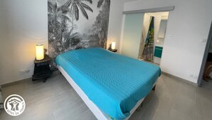 Ein Schlafzimmer mit einem Bett, zwei Nachttischlampen, einem großen tropischen Wandbild und einem durch eine offene Tür einsehbaren Badezimmer.