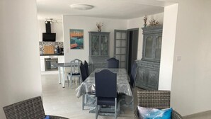 Ein Essbereich mit einem Tisch, gedeckt für vier Personen, umgeben von Korbstühlen mit Kissen. Im Hintergrund ist eine Küche mit einer gefliesten Spritzschutzwand und einem Kühlschrank zu sehen.