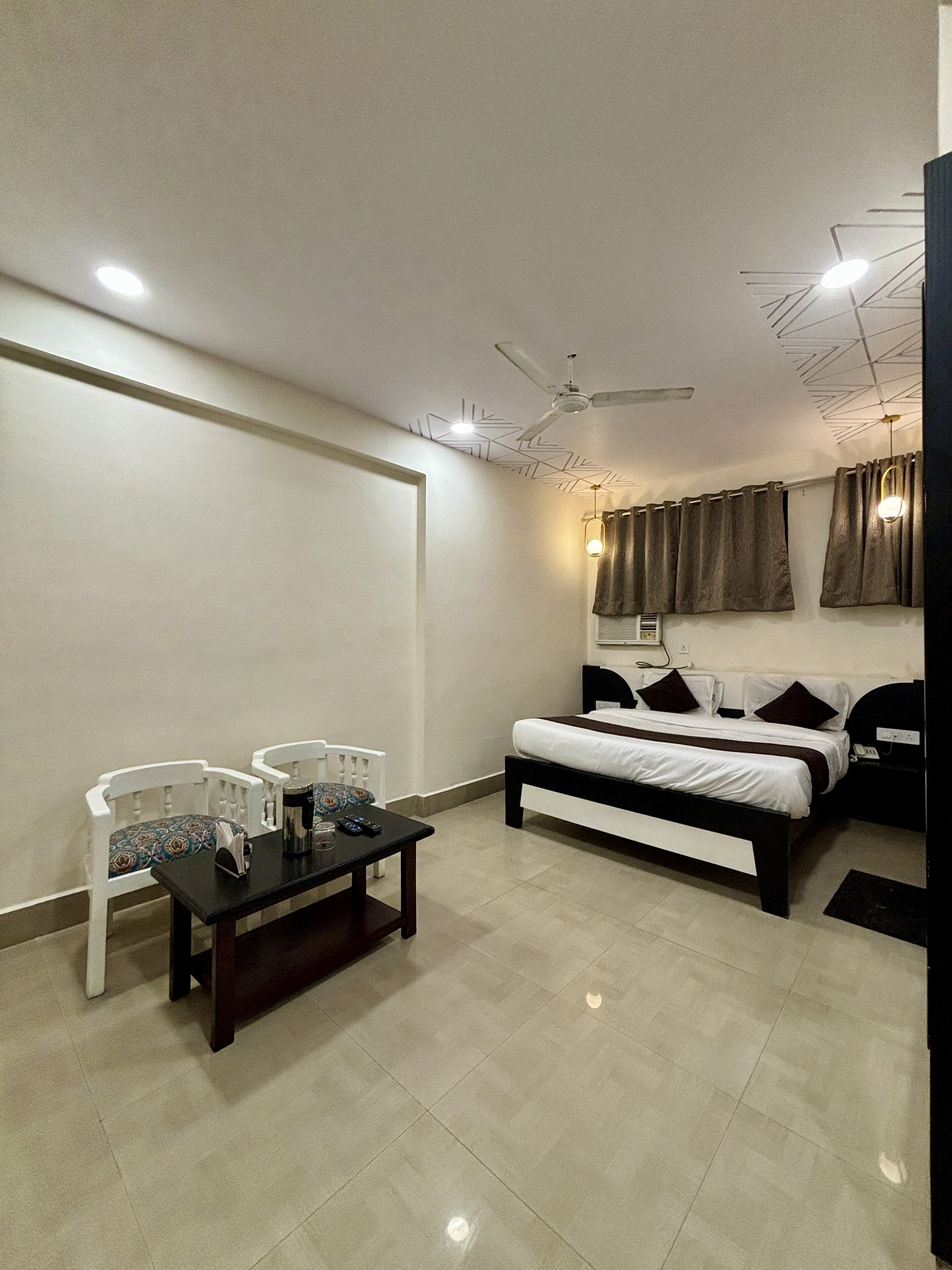 A modern hotel room with a bed, a desk, two chairs, a ceiling fan, and a window with curtains.