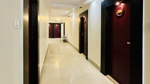 Un pasillo de hotel con piso de baldosas brillantes, puertas de colores oscuros y claros, y un letrero de ascensor visible.