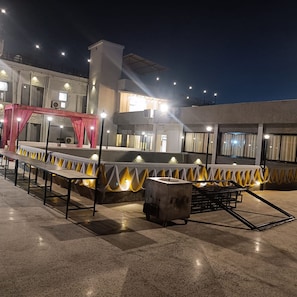 An outdoor seating area with tables and chairs, a stage with red curtains, and a building with multiple windows and lights. - hotel mahamaya (Satna)