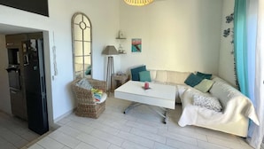 A compact living space with a kitchenette, a white sofa with teal cushions, a wicker chair, and a hanging light fixture.