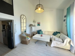 A compact living space with a kitchenette, a white sofa with teal cushions, a wicker chair, and a hanging light fixture.