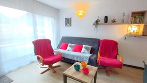 Un salon avec un canapé gris, deux fauteuils rouges, une table basse en bois sur laquelle sont posés des bols colorés, et une grande fenêtre ornée de rideaux légers.