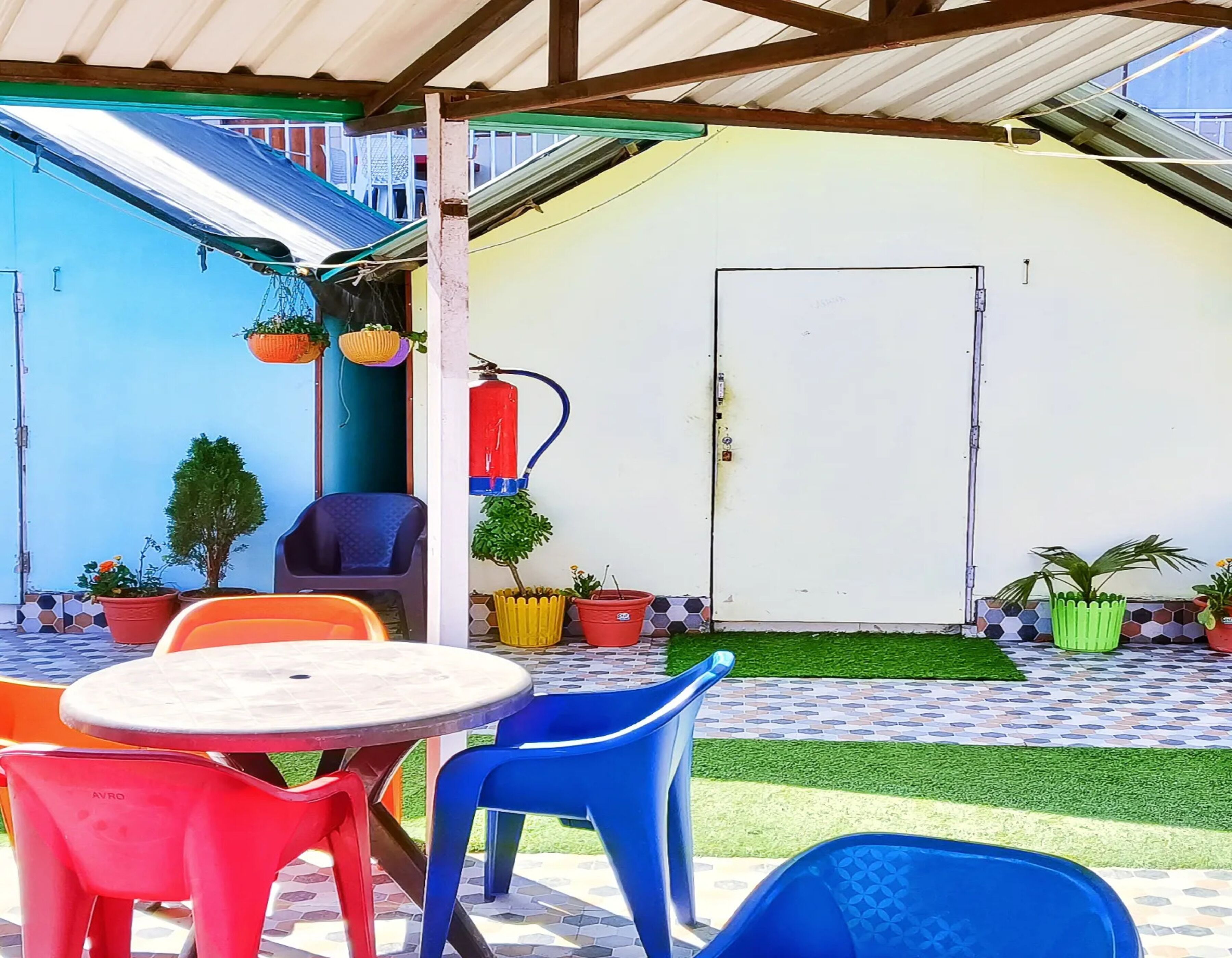 A covered patio area with a round table and blue and red plastic chairs, surrounded by potted plants and a door.