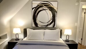 A bedroom with a bed, two bedside tables with lamps, a large abstract painting on the wall, and a sloped ceiling. - The House Hotels - Superior Road (Cleveland Heights)
