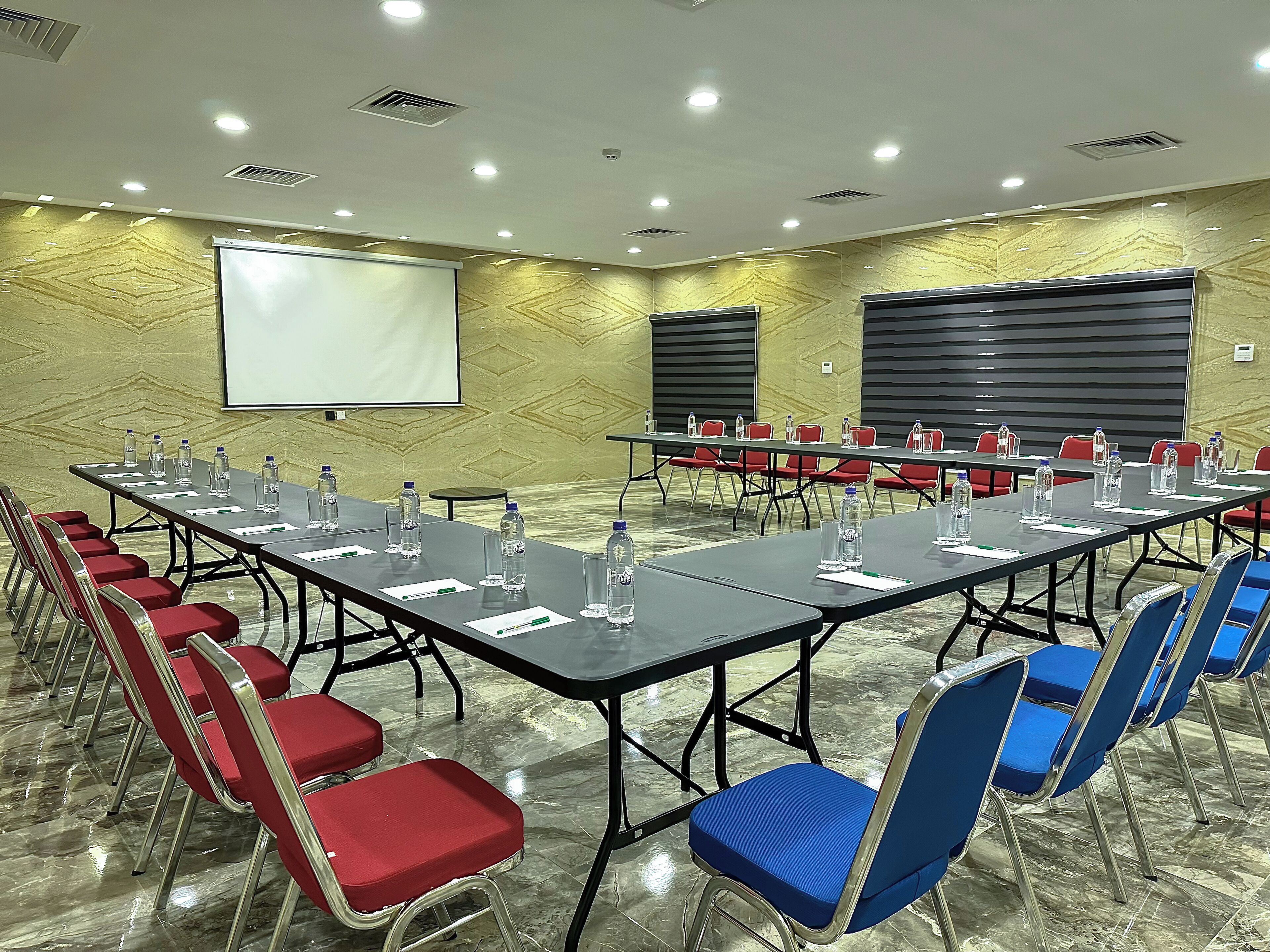 A conference room with a long table, red and blue chairs, water bottles, and a projector screen.